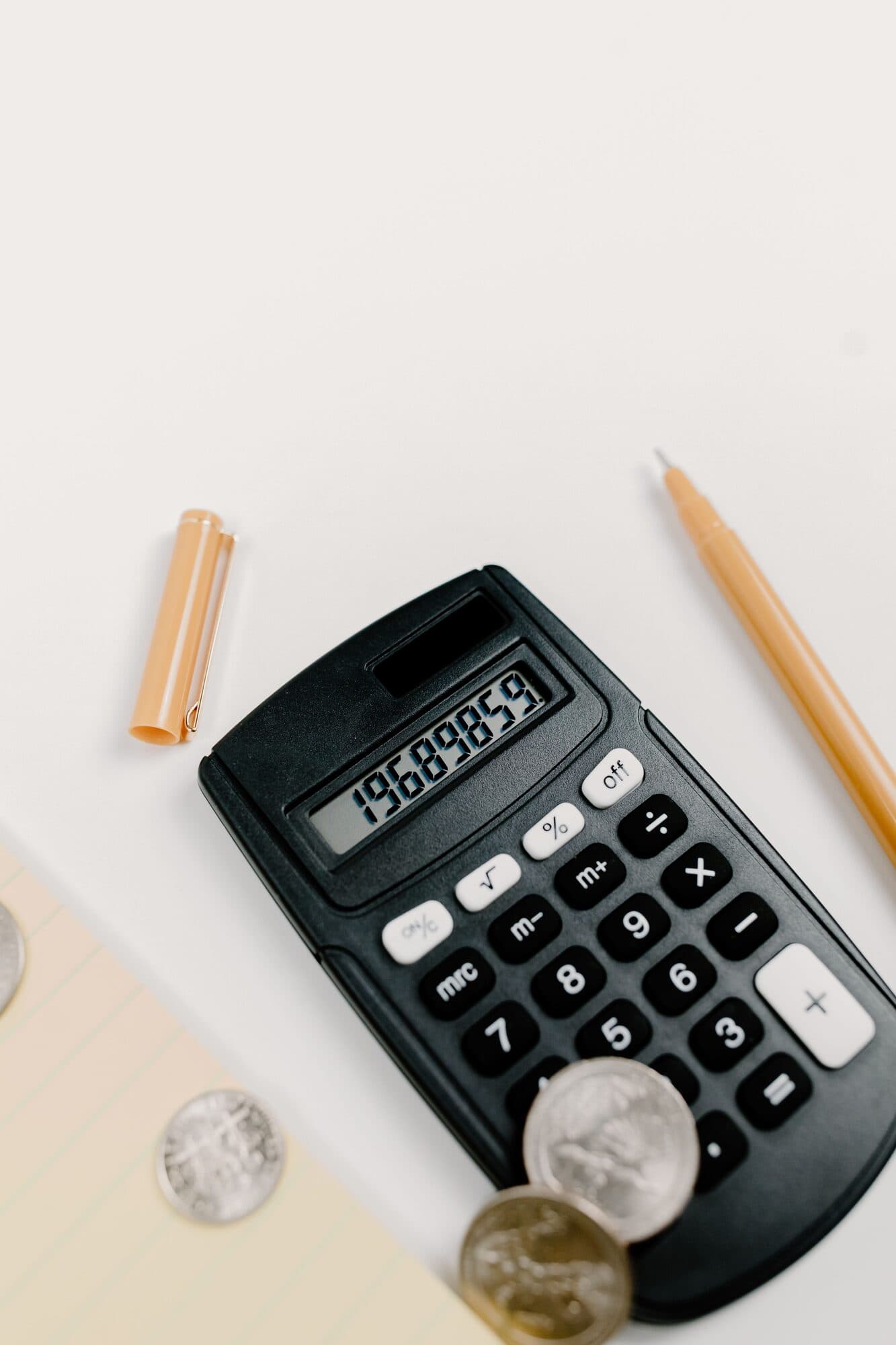 Calculadora sobre uma mesa branca, com caneta, bloco de notas e moedas ao lado